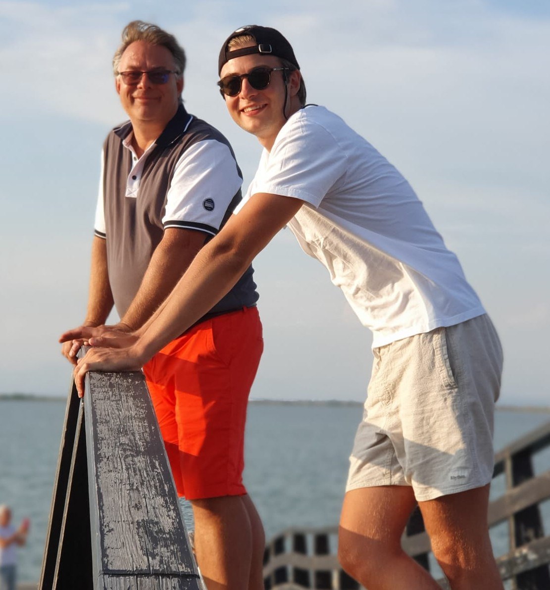 Oliver Fendt und sein Sohn Felix Fendt lächeln gemeinsam auf einer Holzbrücke am Wasser.